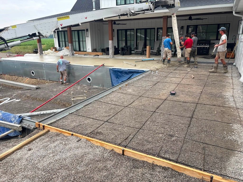 Workers pouring concrete over a gravel base with steel reinforcement around an inground pool