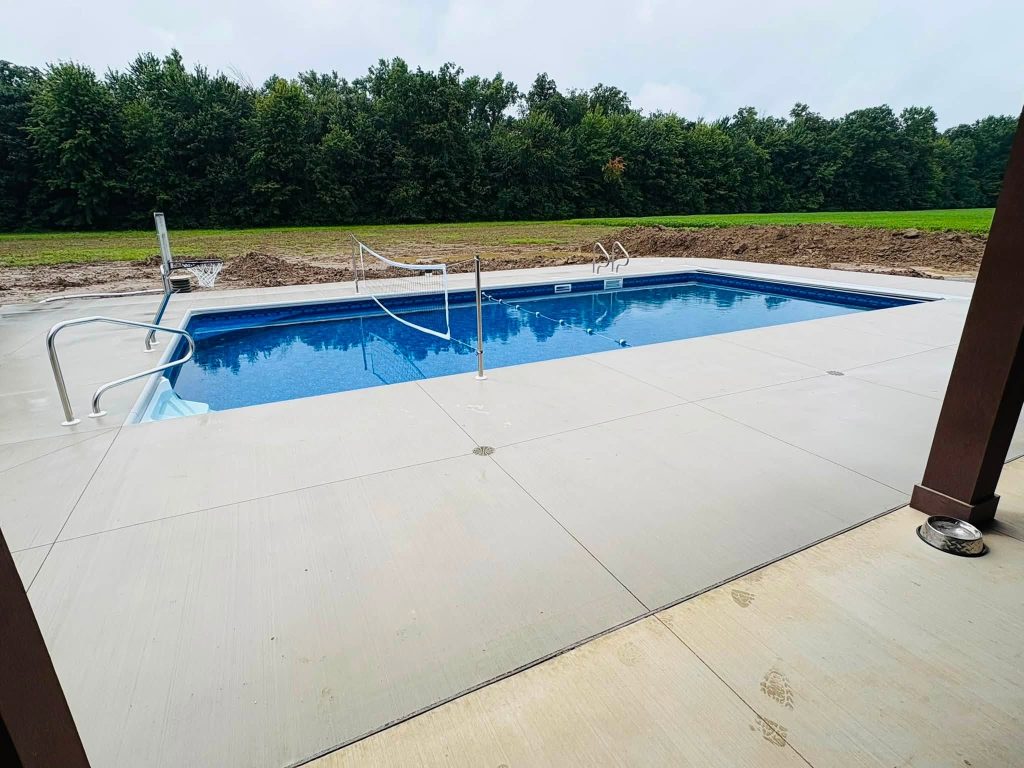 Rectangular inground pool with a concrete deck, entry steps, volleyball net, and basketball hoop
